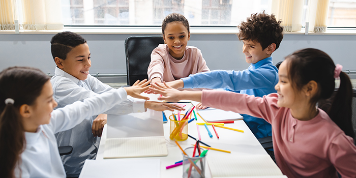  Teamwok And Children Concept. Diverse excited group of emotional happy junior school kids sitting at desk in classroom and putting hands together, having fun, studying or playing game